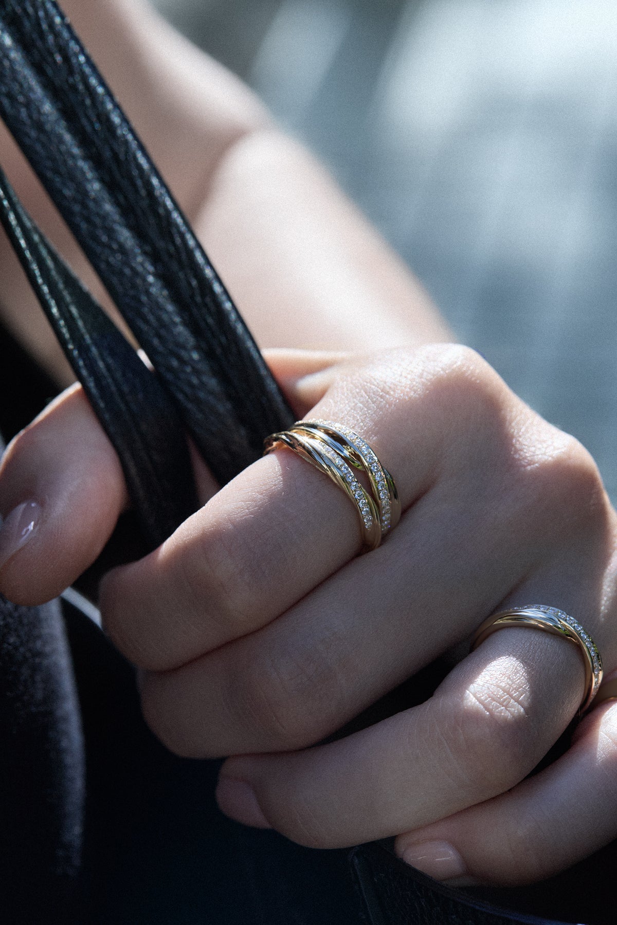 A close-up of a hand wearing three 18 karat gold rings with pavé diamonds and made using Mitsuro hikime, holding a black leather strap.