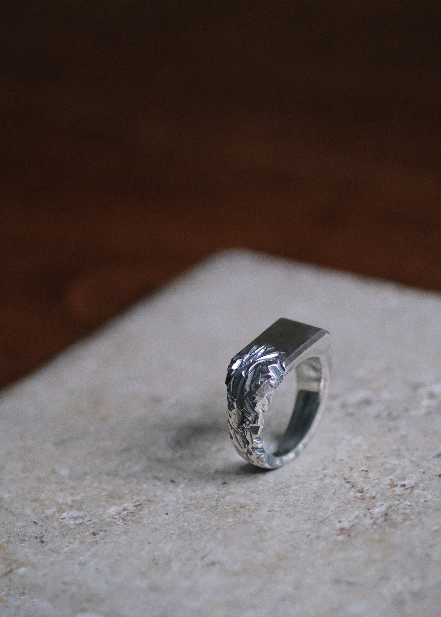 A silver men's ring with a textured, eroded motif on the side, resting on a light beige stone surface with a dark, blurred background.