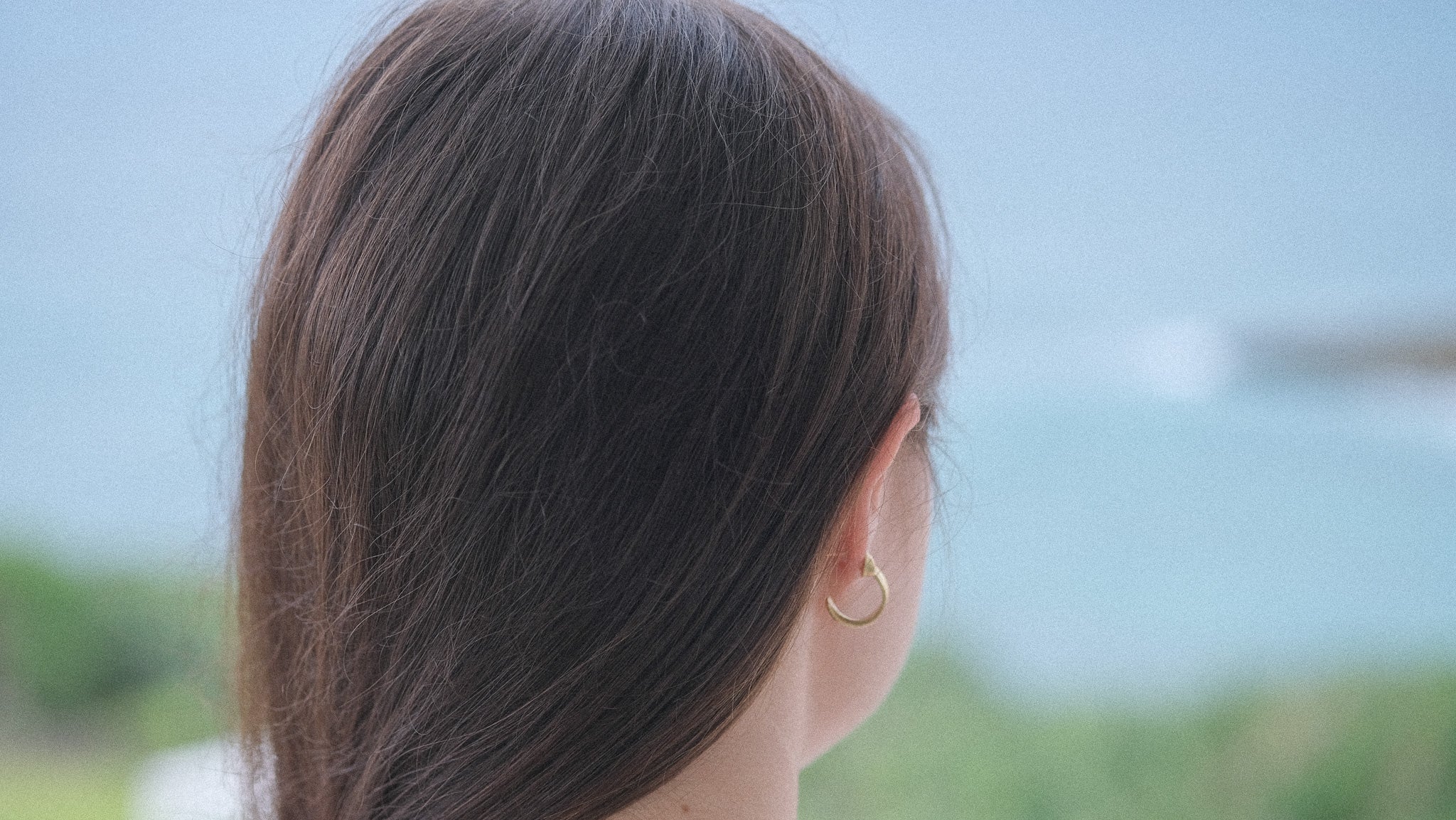 A close-up side profile of a person with long dark brown hair, wearing an 18k gold hoop earring, with a blurred outdoor background of blue sky and greenery.