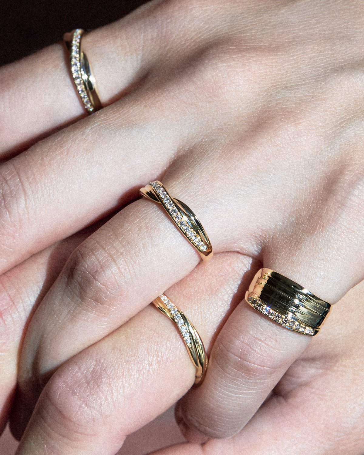 A close-up of hands wearing four gold rings, including three twisted bands with pavé diamonds and one wider bands. All rings feature a Mitsuro hikime texture.