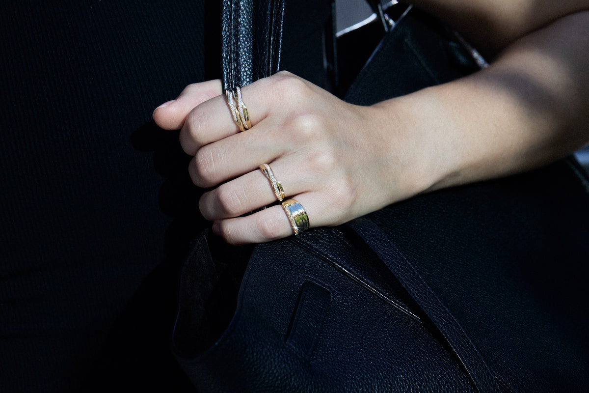 A close-up of a person's hand wearing four gold rings, including two stacked bands and two wider bands, holding onto a black leather bag strap.