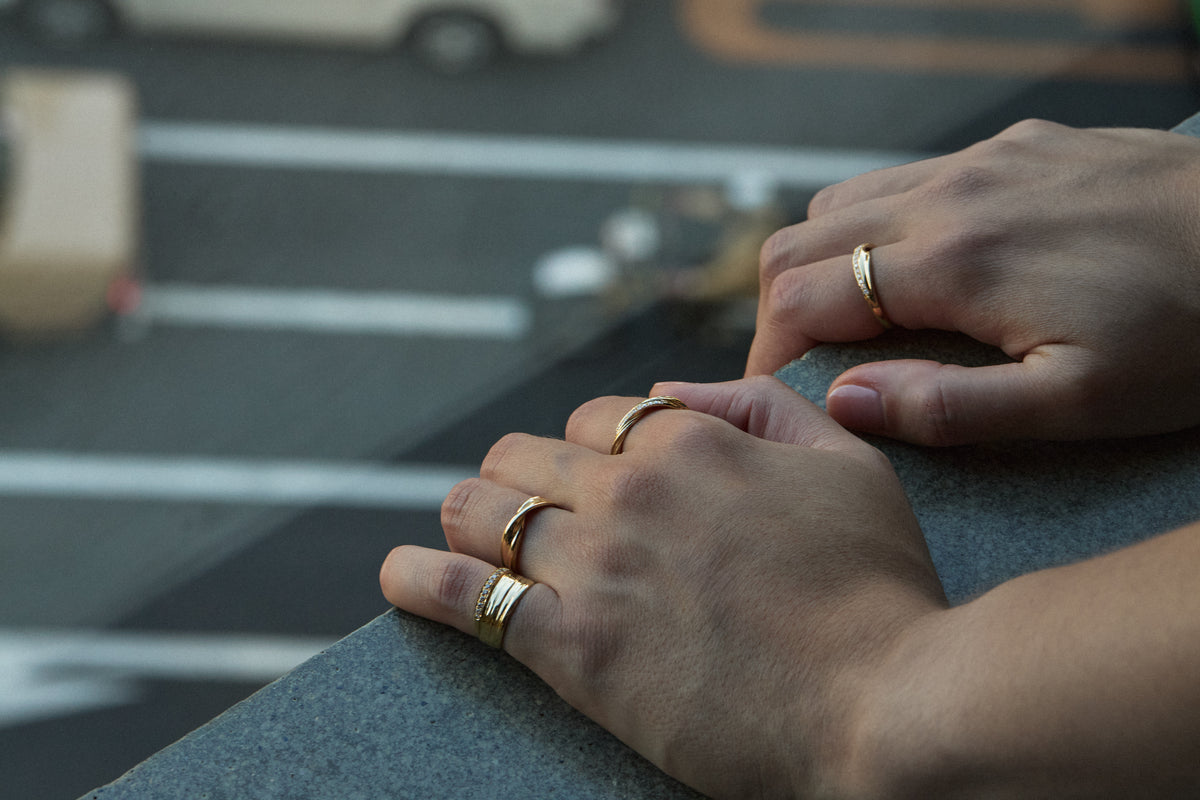 Close-up of a person's hands resting on a gray ledge, wearing several gold Mitsuro hikime rings, with a blurred street scene in the background.