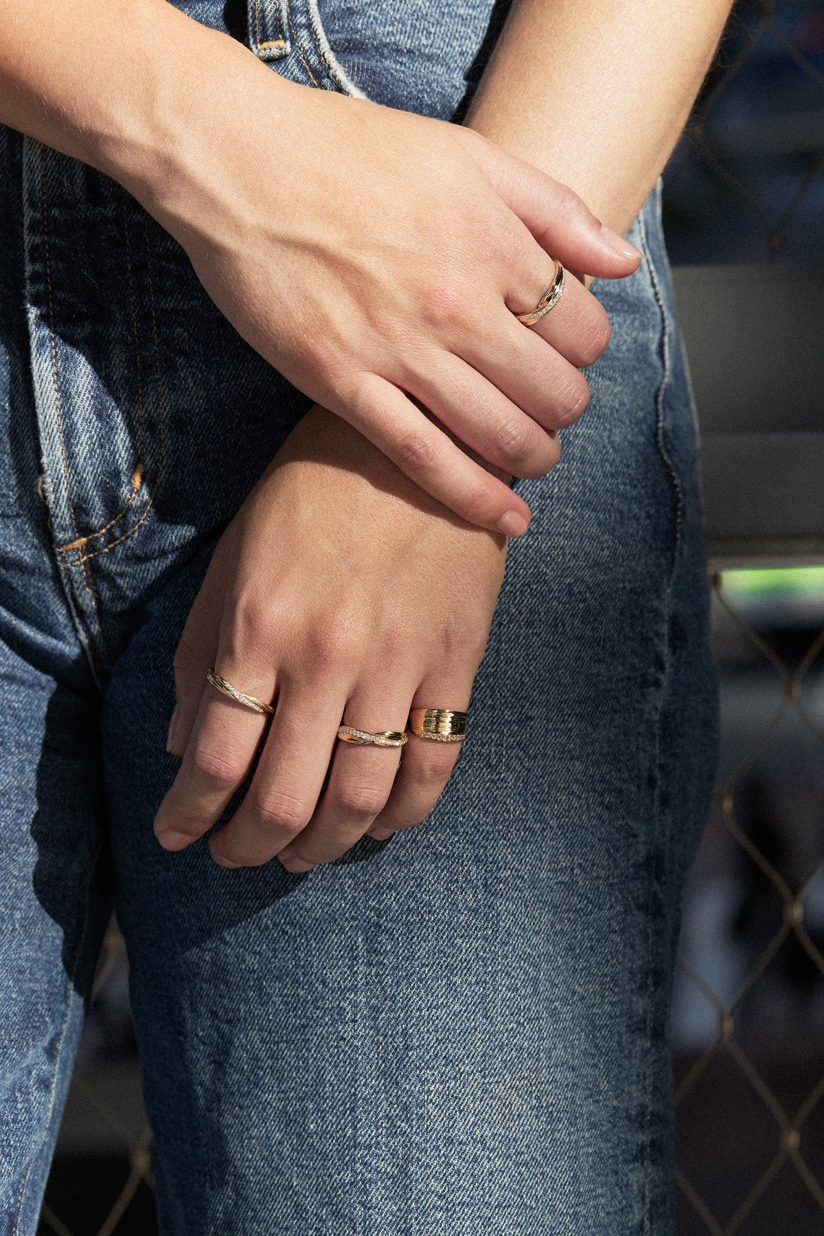 Close-up of a person wearing blue denim jeans and several Mitsuro hikime gold rings on their hands and wrist, with bright natural sunlight and a blurred outdoor background.