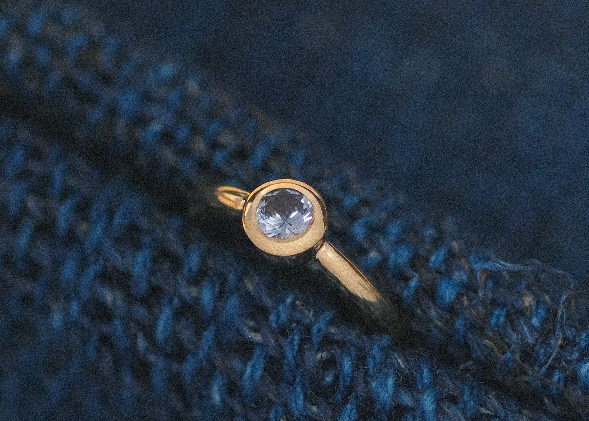 A gold engagement ring with a bezel-set Montana sapphire resting on a textured blue knit fabric background.