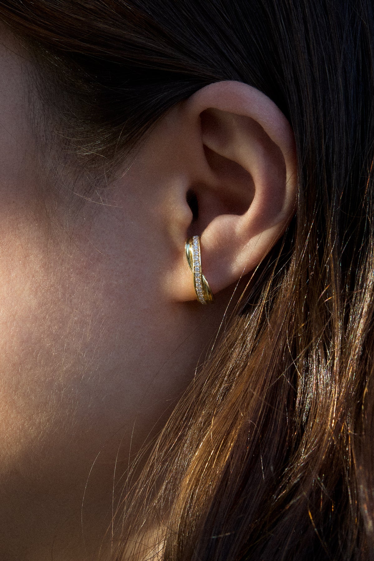A close-up side profile of a woman's ear wearing a small gold huggie hoop earring with a row of pave diamonds, set against her dark brown hair.