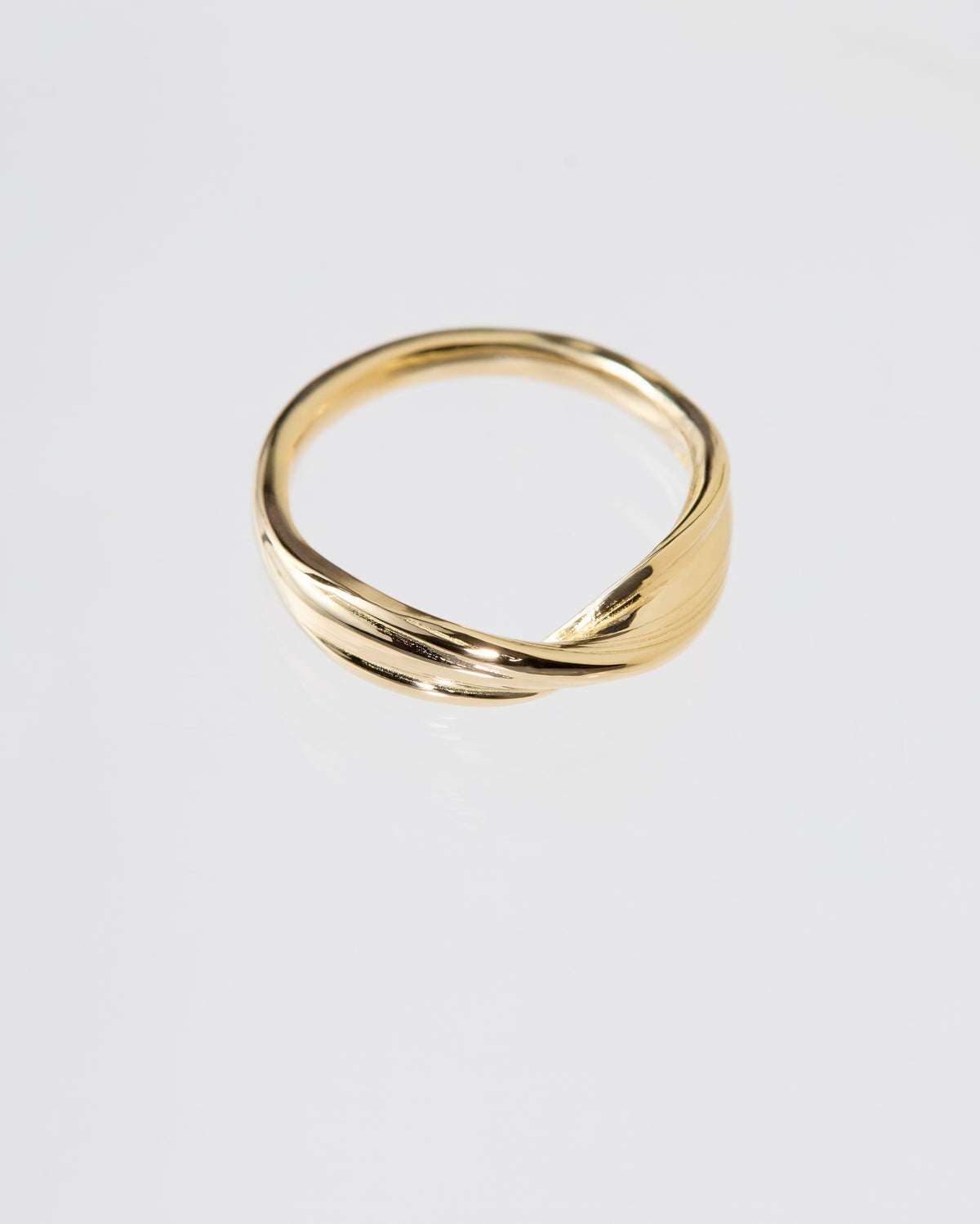A polished gold ring with a twisted, fluid Mitsuro hikime design, photographed on a clean light gray reflective surface.