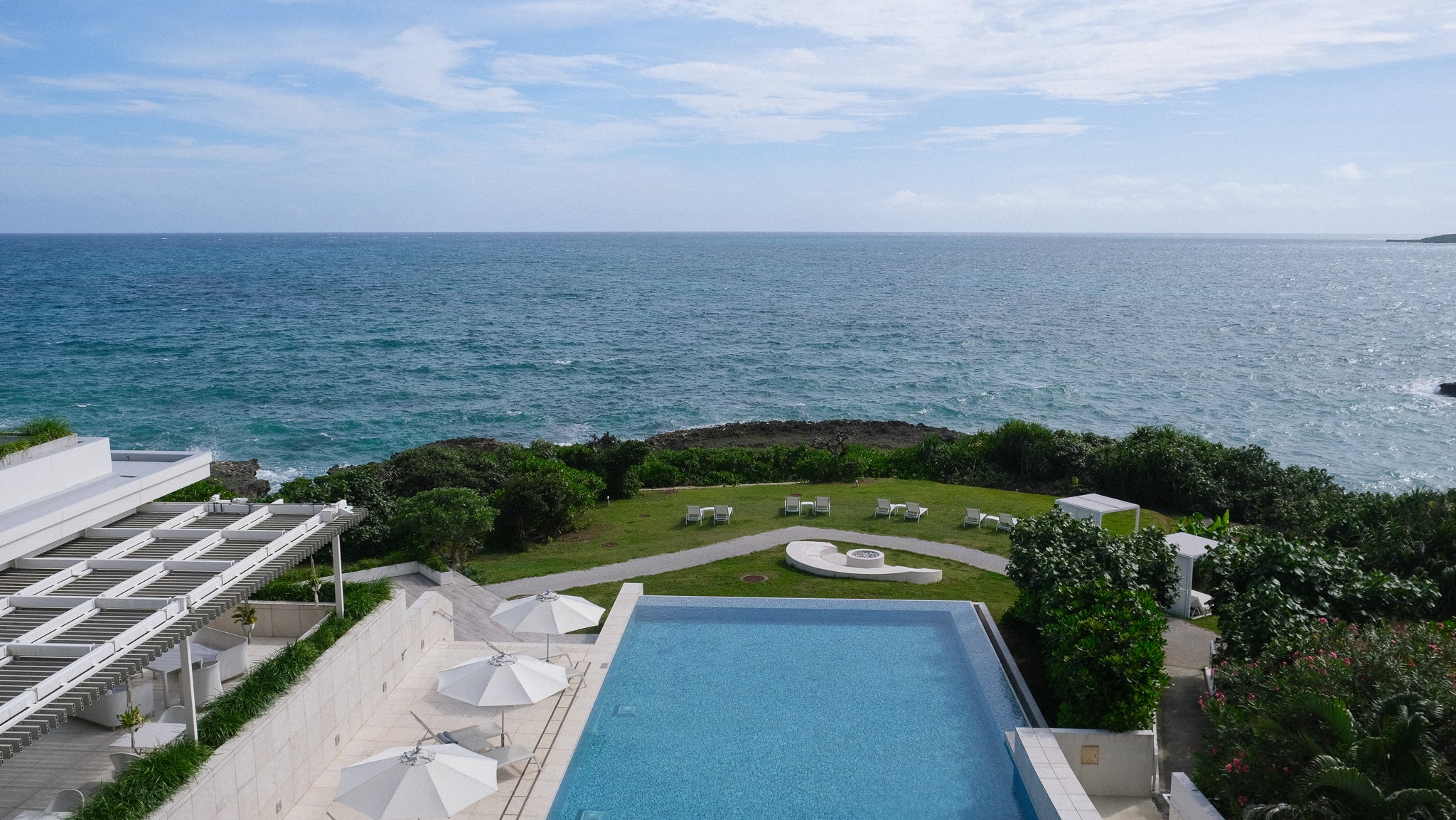 A scenic coastal view with a large ocean expanse under a blue sky, a landscaped green lawn, a rectangular swimming pool in the foreground, and white patio umbrellas beside a building. Iraph Sui Hotel in Miyakojima, Okinawa.