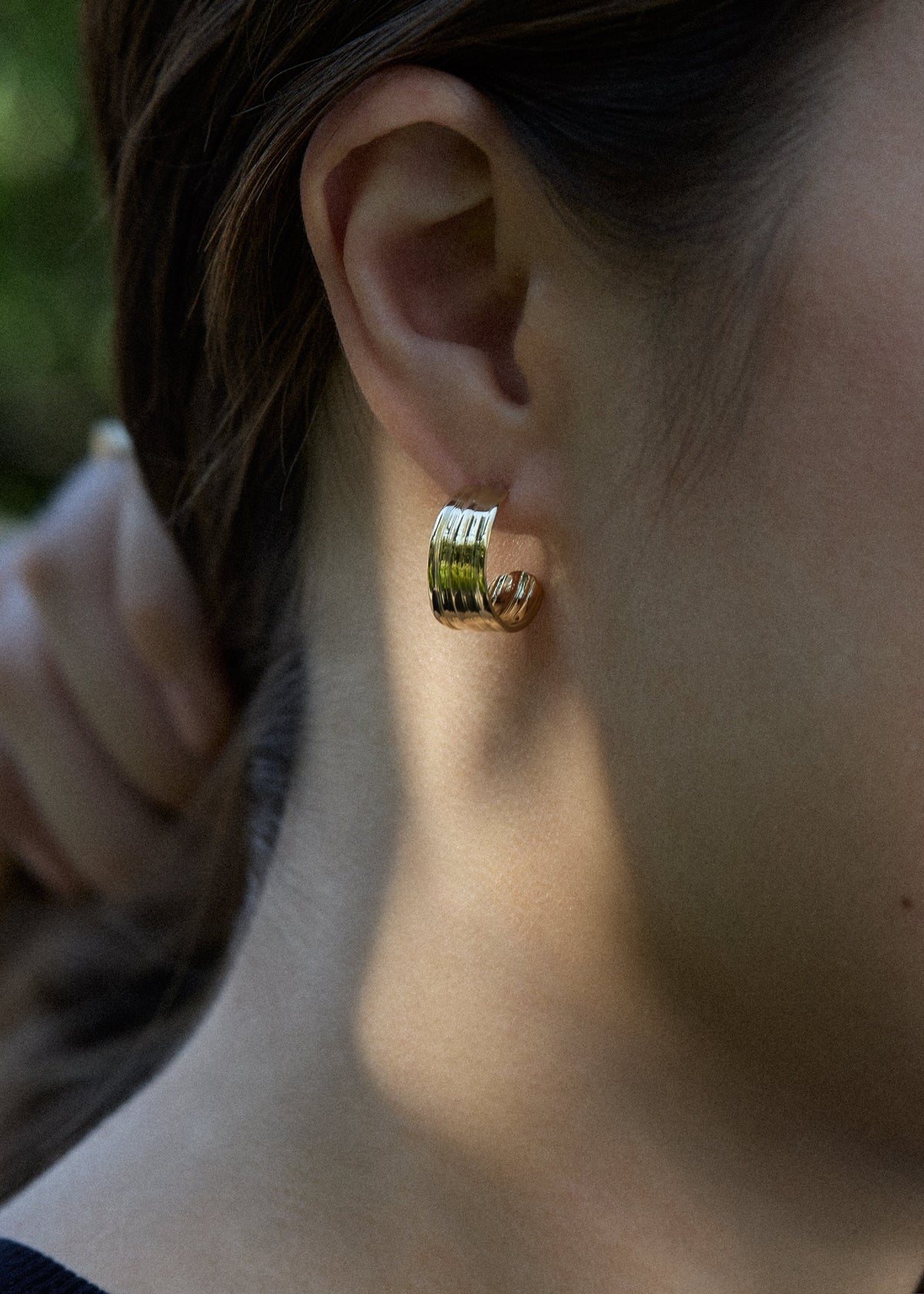 A close-up side profile of a woman wearing a medium gold hoop earring with textured vertical ridges created by Mitsuro wax, set against a blurred green outdoor background.