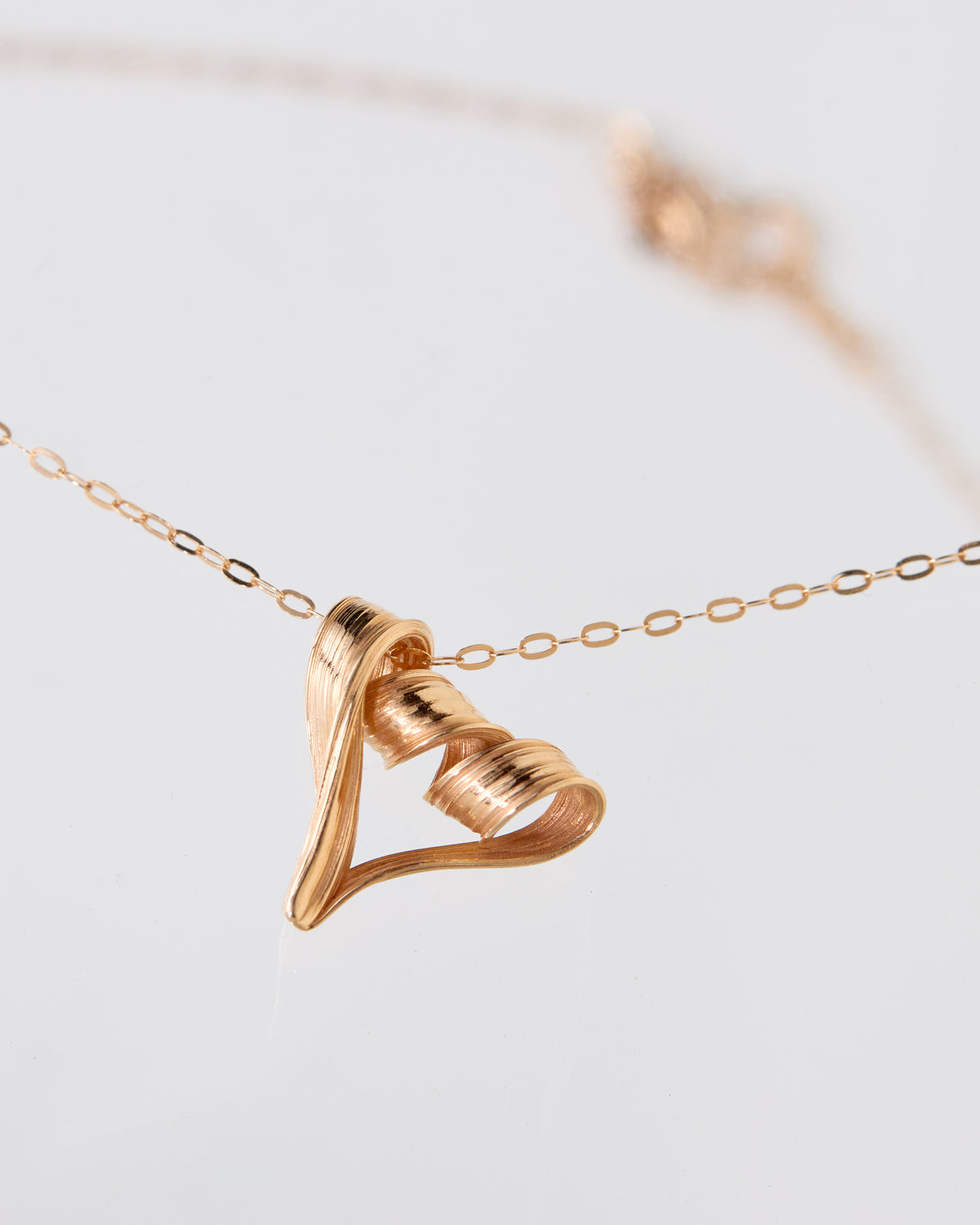 A close-up of a rose gold necklace with a textured, twisted heart-shaped pendant on a delicate chain, set against a soft, light gray background.