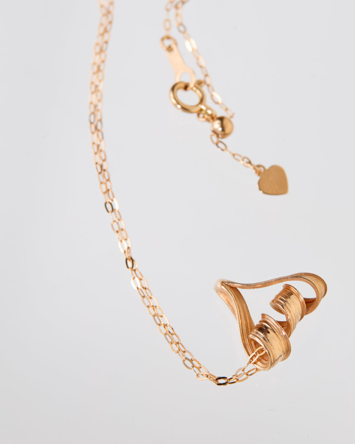 A delicate rose gold chain necklace featuring a large, sculptural twisted heart pendant, displayed on a clean light gray background.