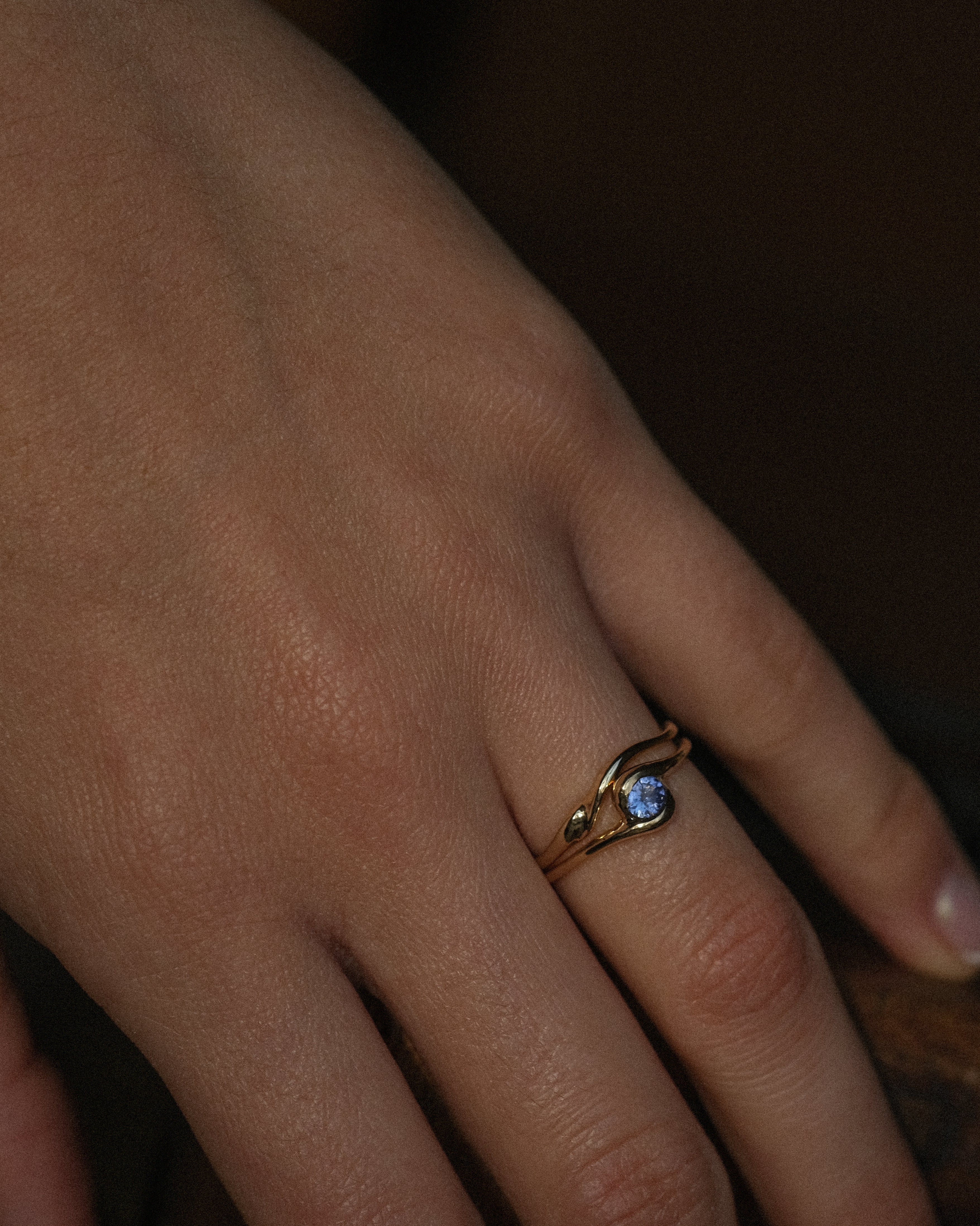Close-up of a hand wearing a gold ring with a blue tanzanite on a dark background