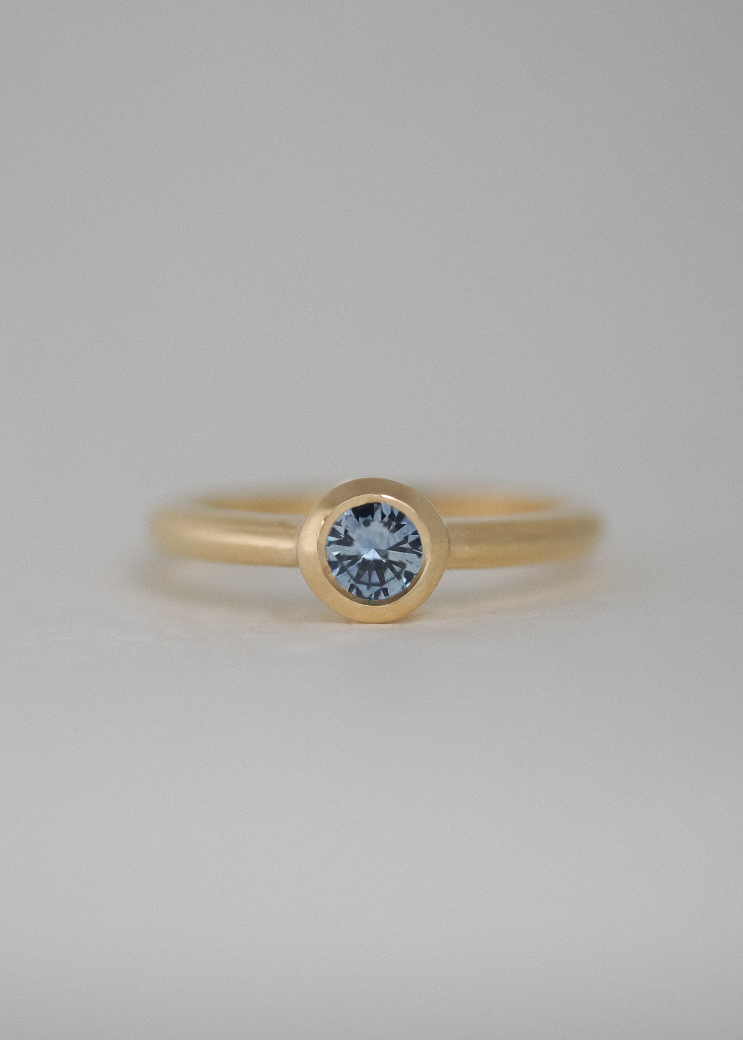 A natural, unheated Montana sapphire in a bezel setting, photographed against a soft, light gray background. A handcrafted and unique engagement ring made in Karuizawa, Japan.