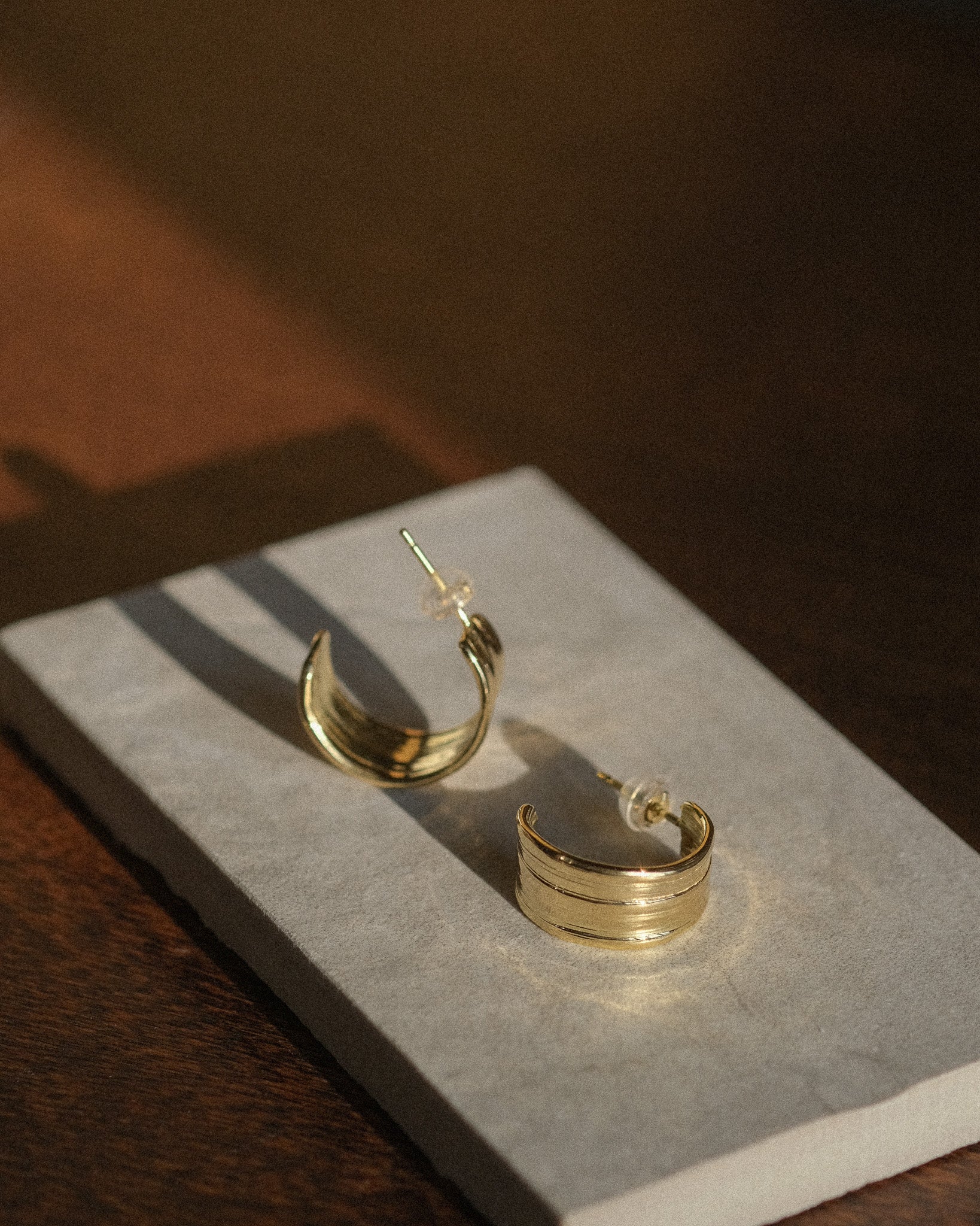 A pair of 18k gold hoop earrings displayed on a rectangular gray stone stand over a dark wooden surface, with warm directional sunlight casting shadows. Made using mitsuro hikime.