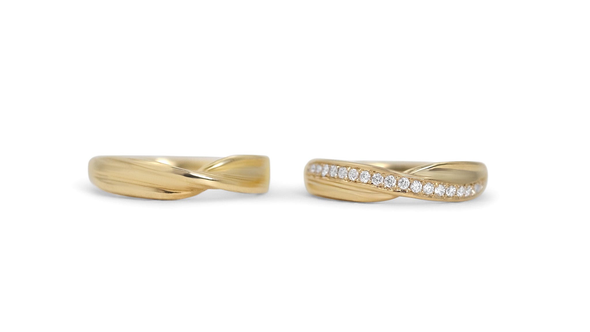A pair of gold wedding band rings on a light gray background. One ring is plain, and the other is embellished with a row of white diamonds.