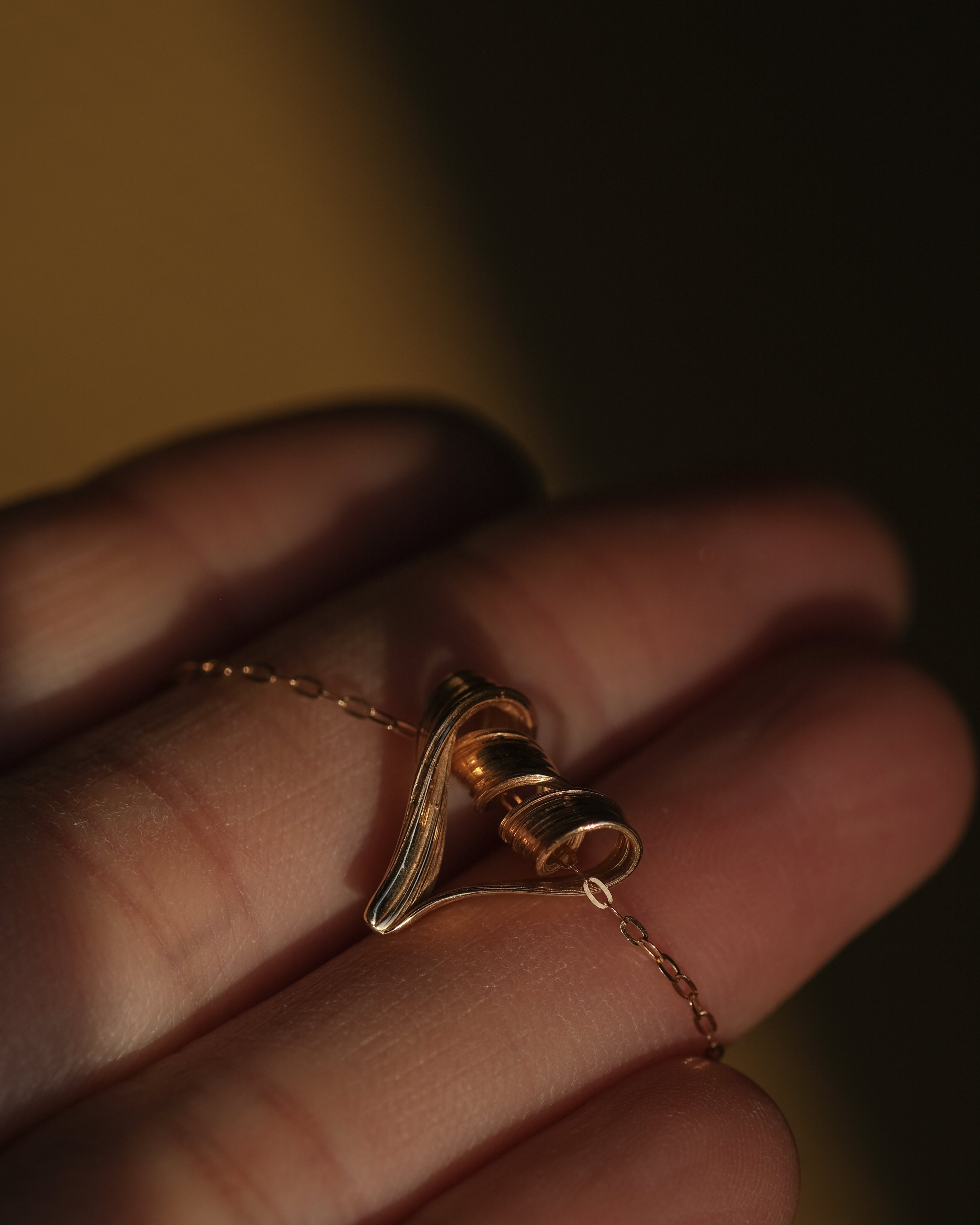 A close-up of a hand holding a delicate gold chain necklace with a small sculptural heart pendant in warm, low-key lighting.