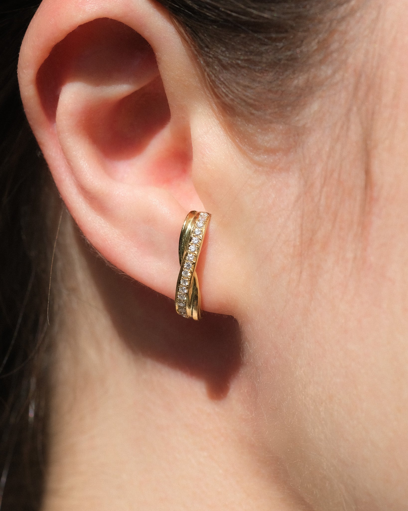 A close-up of a person's ear wearing a twisted gold huggie hoop earring adorned with diamonds on one side.