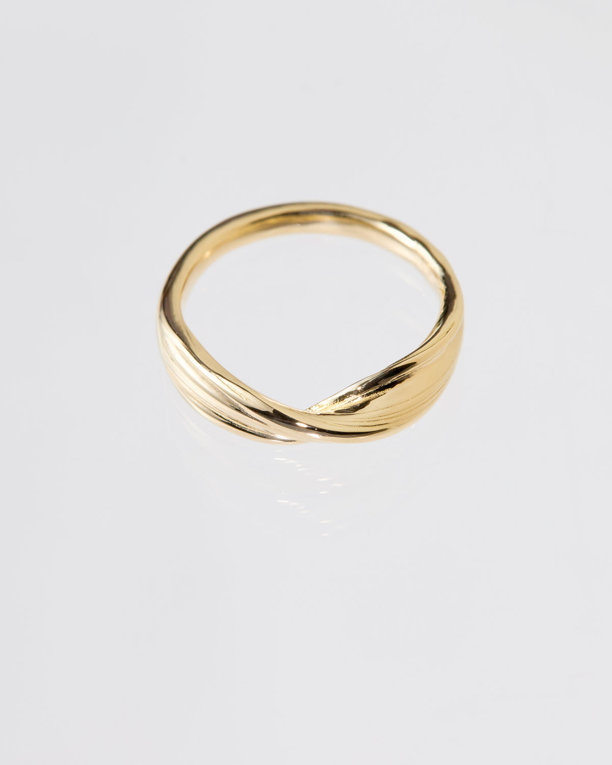 A polished gold ring with a twisted, fluid Mitsuro hikime design, photographed on a clean light gray reflective surface.