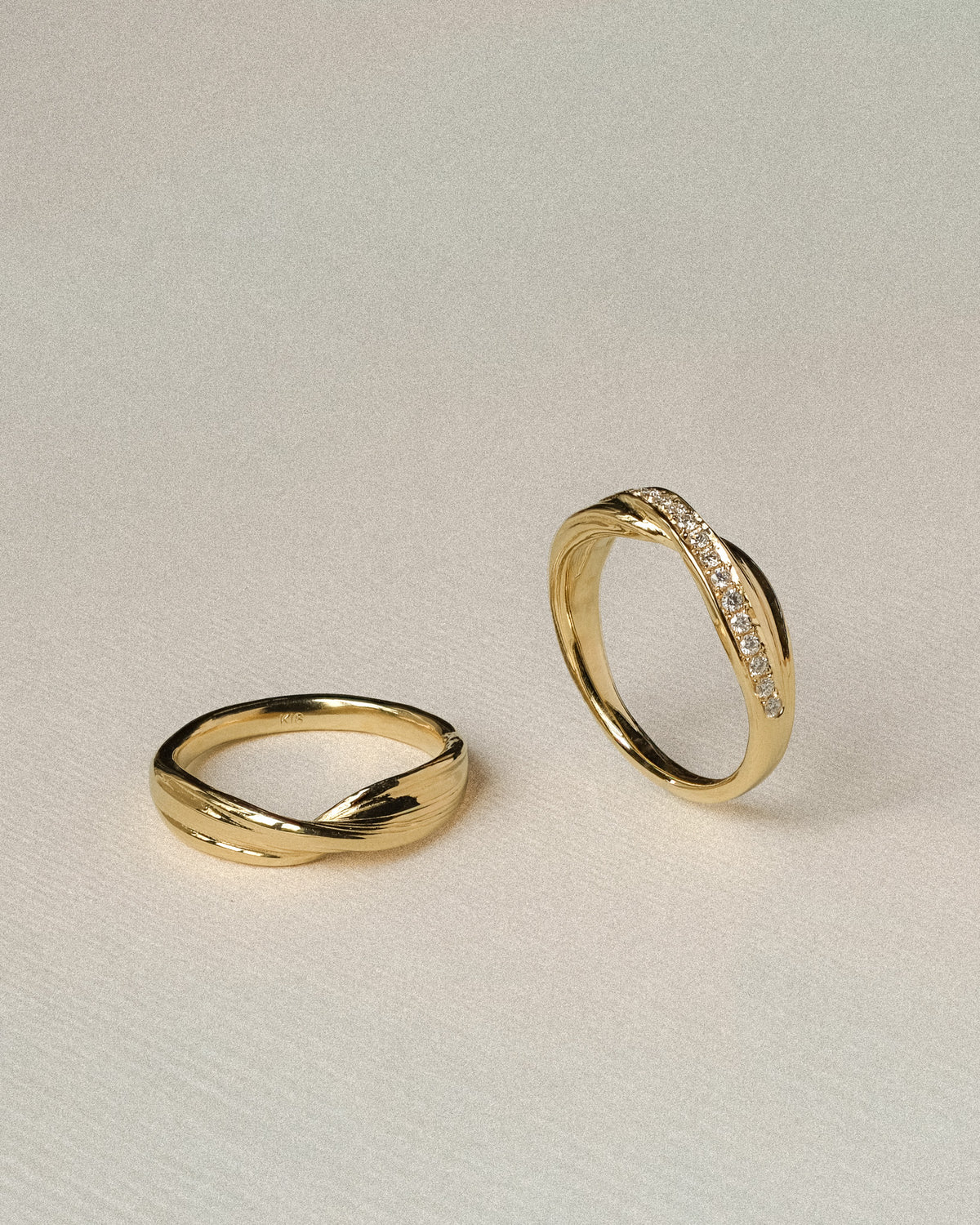 Two twisted gold rings displayed on a light beige textured background. One ring is plain, while the other features a curved row of diamonds along the top.