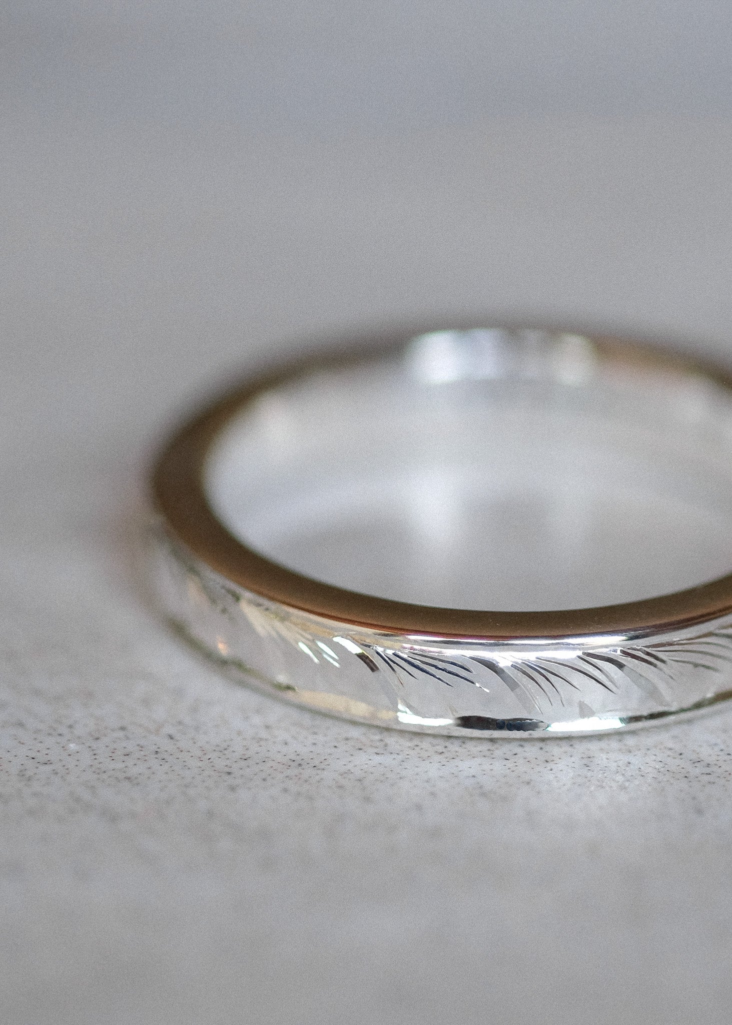A close-up shot of a silver band ring with a hand-engraved. leaf-like pattern on the outer edge and a smooth, polished inner band, resting on a neutral gray surface.