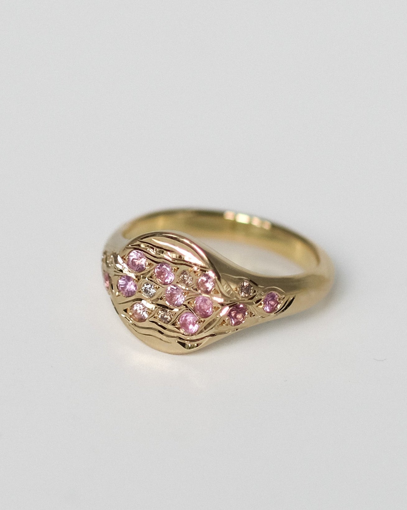 A gold signet ring featuring a wavy, organic-shaped face paved with small pink sapphires and champagne and white diamonds, set against a plain light gray background.