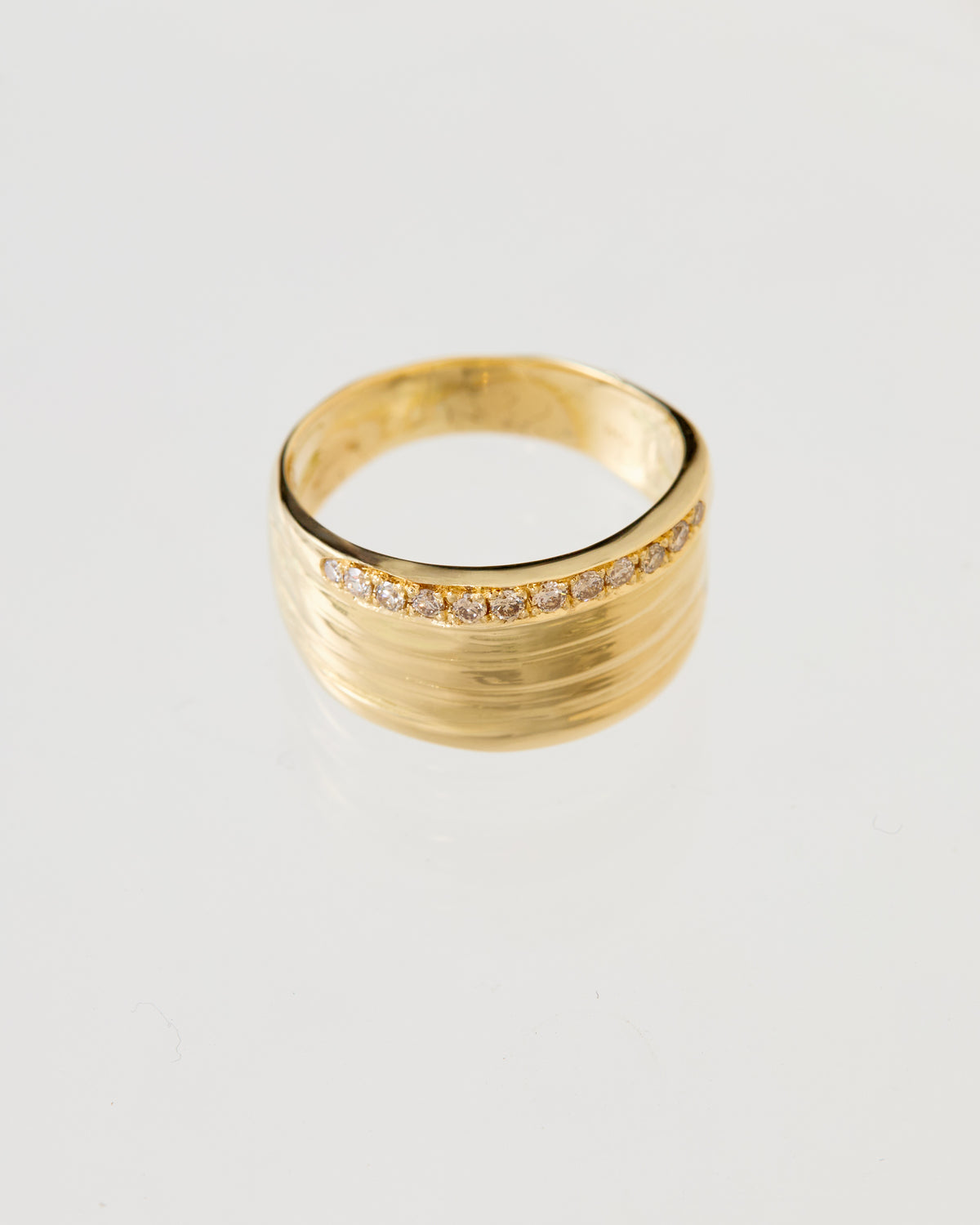 A gold ring with a textured, Mitsuro hikime band and a row of small pavé diamonds along the front, displayed on a reflective white surface.