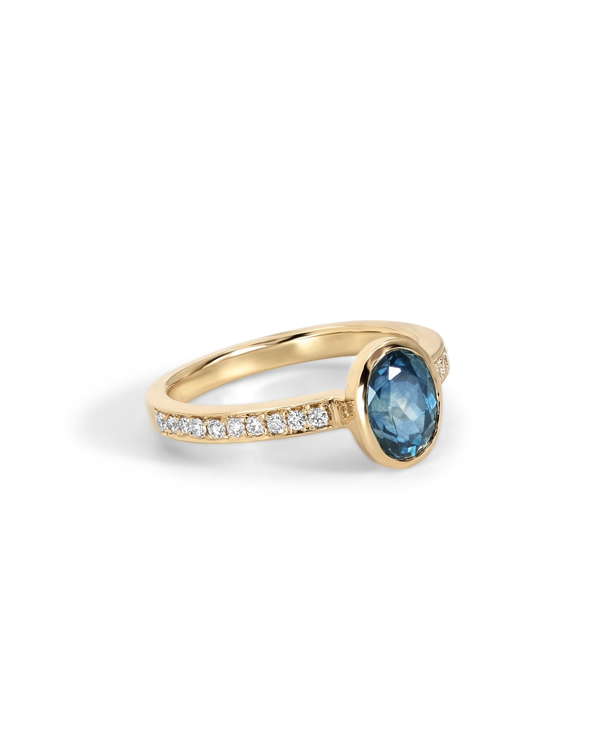 A yellow gold engagement ring featuring a Montana Sapphire in a bezel setting, with a pavé band of diamonds on one side, shown on a plain white background.