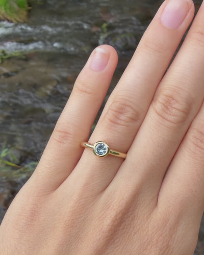 A close-up of a hand wearing a gold ring with a blue Montana sapphire, set against a blurred outdoor background of a river.